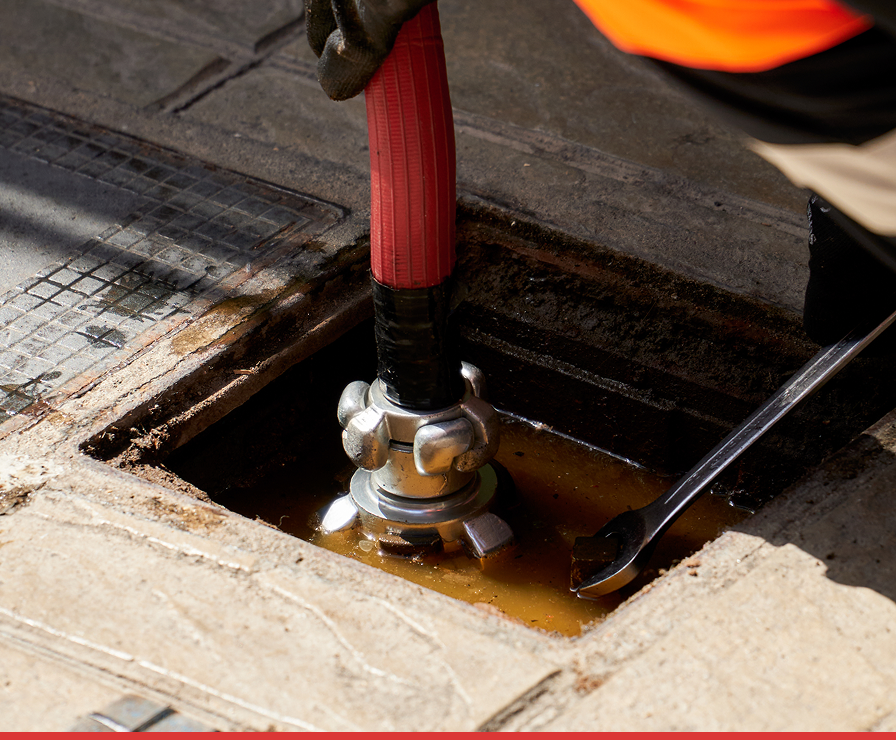 A person wearing an orange sleeve, part of the Divine Restoration Group in Charlotte, inserts a red hose with a metal coupling into a muddy, water-filled utility hole, with a wrench lying nearby on the pavement during property restoration work.
