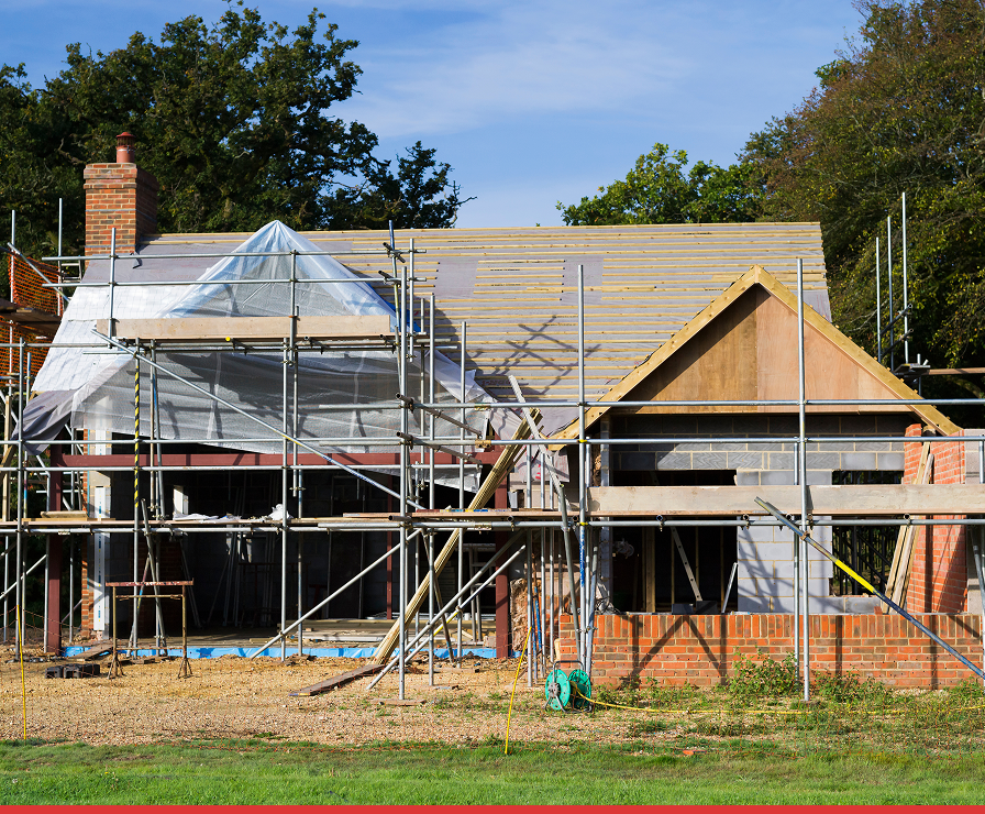 A partially built brick house under construction in Charlotte, with scaffolding set up by Divine Restoration Group. The roof is incomplete, and plastic sheeting covers part of the building. Trees and a blue sky are in the background.