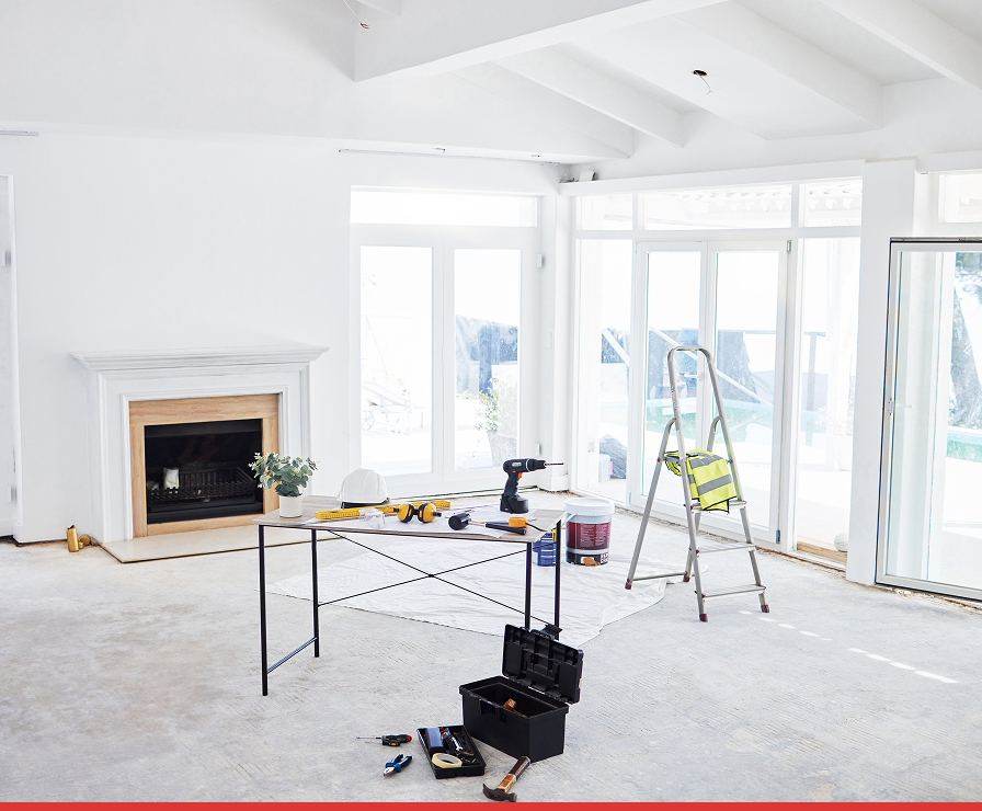 A bright, unfinished room in Charlotte with white walls, a fireplace, and large windows. Renovation work by Divine Restoration Group is underway, with tools and supplies on the table, plus a ladder and open toolbox suggesting property restoration in progress.