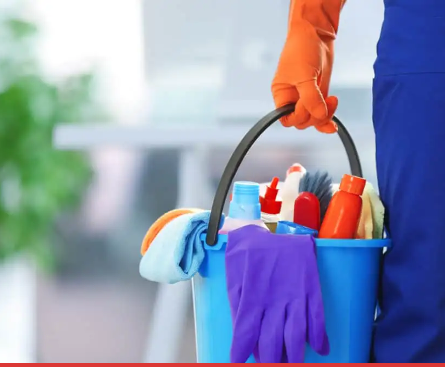 A person wearing an orange glove holds a blue bucket filled with cleaning supplies and purple gloves, ready for Property Restoration. The blurred indoor setting hints at a Charlotte home serviced by Divine Restoration Group.